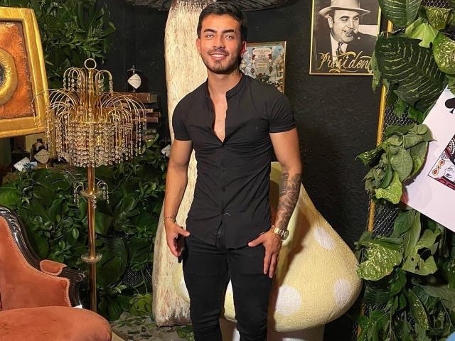 Zhion, muscular, tan-skinned, short brown hair, black shirt, black pants, tattoo on right arm, smiling, standing, holding sides, elegant chandelier, leafy plants, vintage chair.