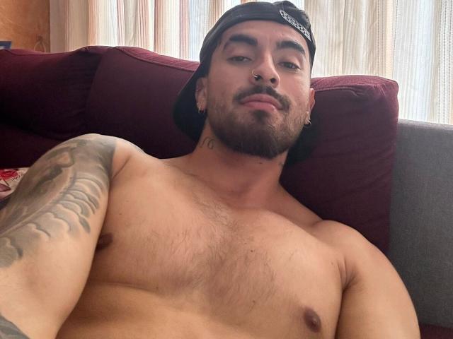 Zhion, sem camisa, com um boné preto virado para trás, tatuagens no braço direito e um piercing no nariz, apoiado em um travesseiro vermelho, tem uma barba rala e pelos no peito..