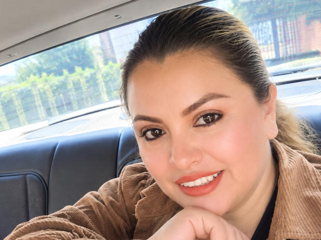 CharlotteDevereaux has brown eyes, full lips with red lipstick, and is smiling in a car, wearing a brown ribbed shirt, with her hand under her chin, looking cute.