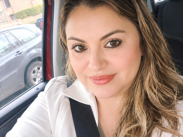 CharlotteDevereaux with wavy brown hair, brown eyes, and pink lipstick, wearing a white shirt, seated in a car with a black seatbelt, smiling subtly. Boobs are not visible.