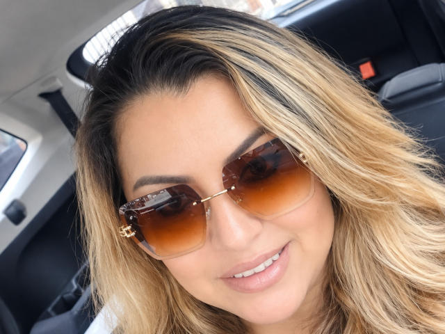 CharlotteDevereaux with blonde, wavy hair, wearing brown, gradient sunglasses and smiling, showing white teeth, inside a car with black seats. Her skin is light brown and she's cute.