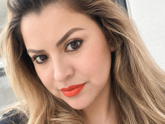 CharlotteDevereaux has bright orange lipstick, brown eyes, and blonde wavy hair. Her eyebrows are thick and she's smiling slightly with a cute face.