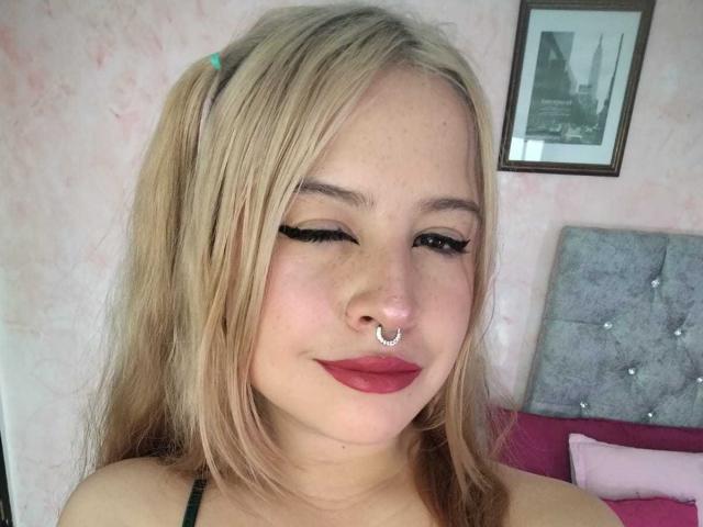 MelaniBecker with blonde hair, nose ring, and red lipstick, winking with black eyeliner, smiling, wearing green top, head against grey tufted bed, pink pillow, framed picture above.