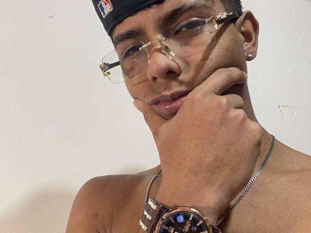 KingFastt with glasses, black cap, earrings, and a brown wristwatch, touching his chin with a slightly smug expression, shirtless, and wearing a silver chain.
