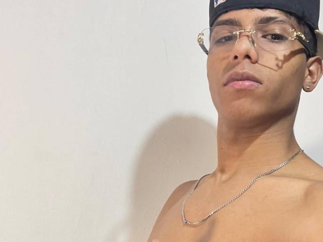 KingFastt, shirtless, with a chain necklace, wearing glasses and a black cap, has a serious face, brown skin, and a slight shadow on his face.