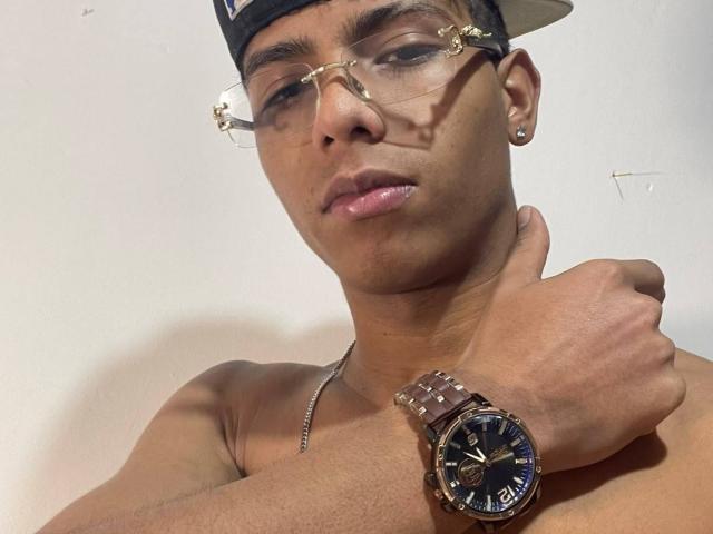 KingFastt with glasses, a cap, and a wristwatch, holding his neck, shirtless, brown skin, slightly pouty lips, and a small ear piercing on the left side.