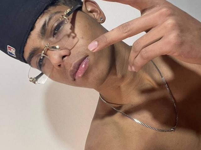 KingFastt with glasses, black cap, and chain, pointing at nose with index finger, shirtless, showing a clean-shaven face, and a small ear piercing, looking serious.