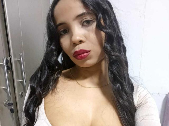 HotAngelica69 has long, wavy black hair, red lipstick, and a gold necklace. She's wearing a white, low-cut top that shows her boobs. She's looking at the camera.