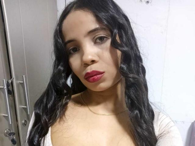HotAngelica69 with wavy black hair, red lipstick, and a low-cut white top, showing her tits and a gold necklace. She's looking directly at the camera, with a neutral expression.