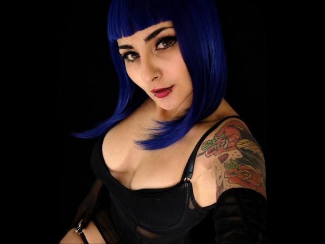 Carll with blue bob, black dress, showing off her tits and colorful tattoo on her right arm, black background, looking up with red lips and a nose ring.