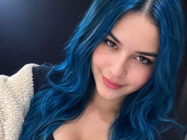 AuroraSantorini has blue hair, brown eyes, and a cute smile. She's wearing a fluffy white jacket, showing her tits slightly with a black top. She's a pretty girl.