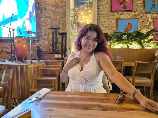 RousseColliinss has red hair, glasses, and is smiling while wearing a white dress with a floral button, sitting at a wooden table with her hand on her tits.