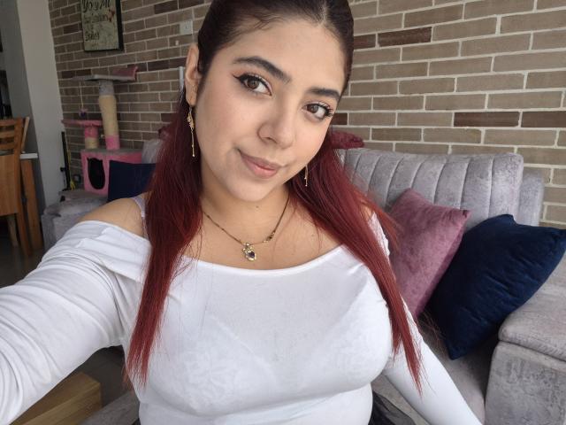 RousseColliinss has red hair, white off-shoulder top showing her tits, subtle makeup, and a confident smile, sitting on a grey couch with purple and navy pillows.