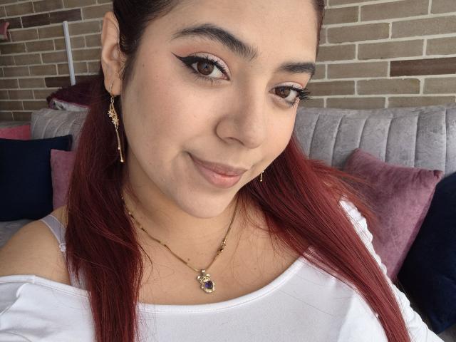RousseColliinss has red hair, brown eyes, and wears a white top with gold earrings and necklace. She's smiling with winged eyeliner, showing off her cute face.