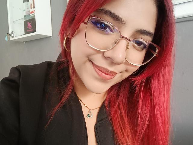 RousseColliinss with red hair, glasses, and hoop earrings, smiling with pink lips, wearing a black top, and a green pendant. Her blue eye makeup stands out.