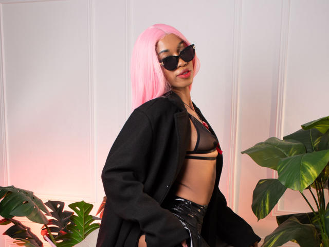 AllisonRomano with pink hair, black sunglasses, sheer black bra, and black leather pants, wearing an open black coat, standing next to green plants, showing her tits.