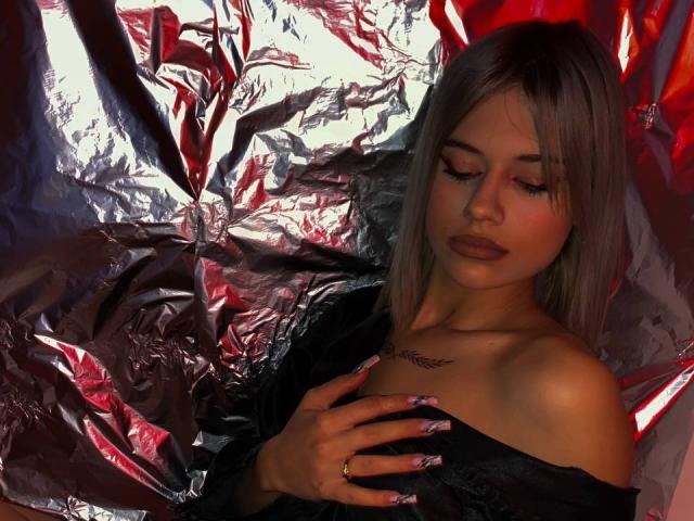 IvyVeil with silver hair, dark lipstick, and long nails covering her tits with a black shirt, hand with ring, tattoo on chest, looking down, silver and red foil background.