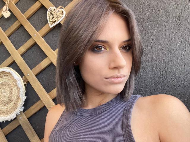 IvyVeil, con i capelli grigio-marrone che le arrivano alle spalle, occhi marroni e un trucco leggero, indossa una canotta a coste viola, mostra un'espressione neutra ed è in piedi di fronte a una parete scura e strutturata..