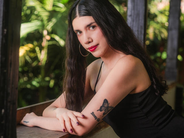 MaddyMedoza met lang zwart haar, rode lippenstift, oorringen, een zwart tanktopje, een getatoeëerde arm, leunt tegen een houten raam, groene begroeiing buiten, kijkt naar de camera, paarse nagellak..