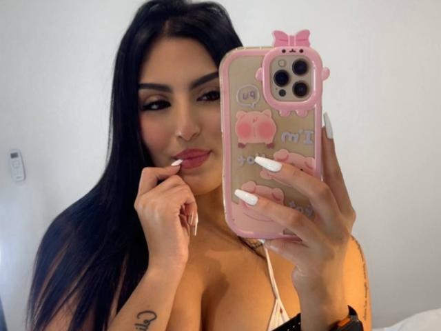 AriiaMon with long black hair, holding a pink phone case with cute designs, biting her white-manicured finger, showing large tits, and a small tattoo on her hand.