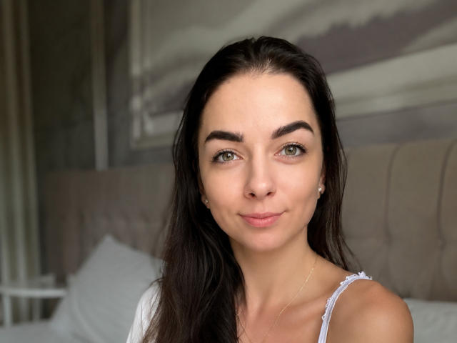 PolinkaN has a cute face with green eyes, dark brown hair, and a small nose, wearing a white top, with a subtle smile, and a delicate gold necklace.