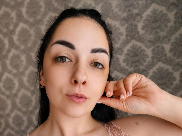 PolinkaN's face with dark brown hair, green eyes, and pink lips, touching her ear with her right hand, wearing a pink lace top, looking forward.
