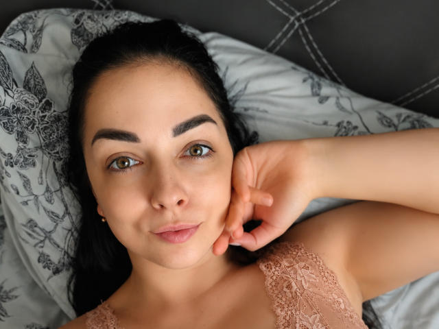 PolinkaN has light brown eyes, black eyebrows, and a slight smile, lying on a bed with lace-covered tits, her hand near her face, and black hair.