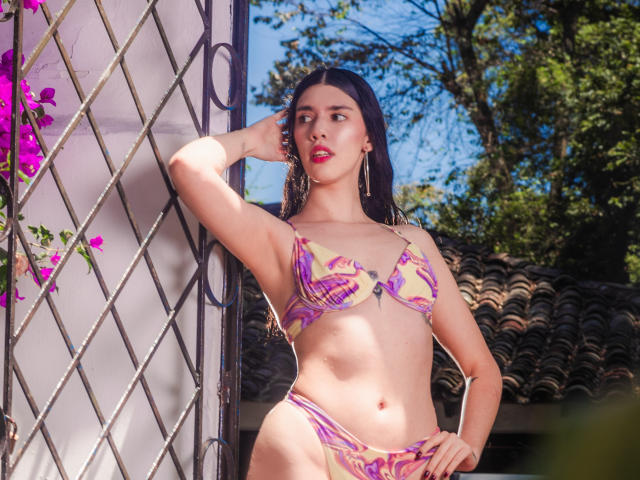 MaddyMedoza mit langen schwarzen Haaren, rotem Lippenstift und einem violetten Bikini, steht vor einem Metallgitter, eine Hand auf dem Kopf, blickt zur Seite und zeigt ihre Brüste und ihren Hintern..