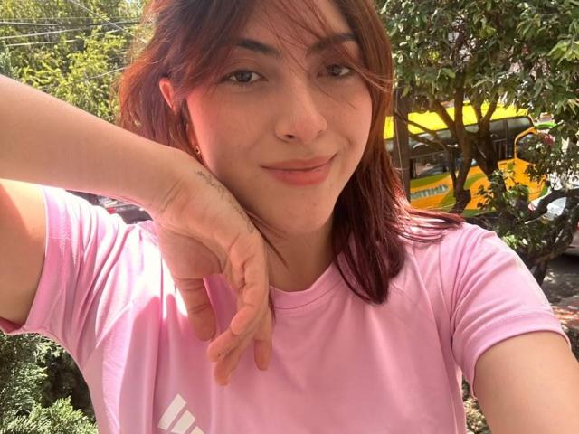 SpencerLe with red-brown hair, light skin, and a subtle smile, wearing a pink shirt, rests her chin on her hand, showing a tattoo on her wrist.