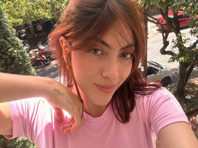 SpencerLe with red hair, pink shirt, and tattoo on left wrist, smiling at camera, holding neck, brown eyes, subtle makeup, outdoor sunlight, visible car and tree in background.