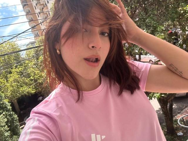 SpencerLe with dirty brown hair, pink t-shirt, tongue out, hand in hair, small earrings, tattoo on right arm, light skin, green leafy background, sunny day, blue sky.