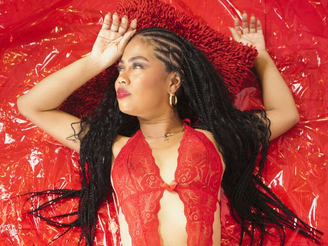 LuciaVargasX with long black braids, wearing red lace lingerie, hands raised, lying on red plastic, red pillow, gold hoop earrings, red lipstick, looking up, arms above head.