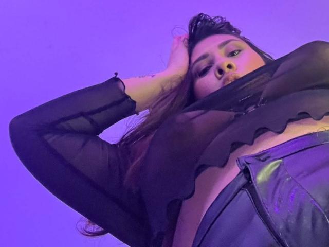 ElyRoss with long brown hair, wearing a sheer black top, has her hand on her head, looking down, with visible breasts and a purple-tinged background.