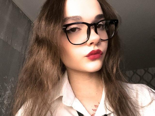 DennyMost has wavy brown hair, black glasses, red lipstick, and a white shirt. She's looking at the camera with a slight pout, showing a tattoo on her neck.