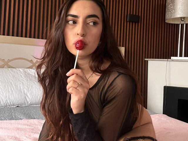 CelesteLarson with long brown hair, sucking red lollipop, wearing sheer black top, exposing tits, pink bed, wooden wall, lamp in background, looking seductively at camera.