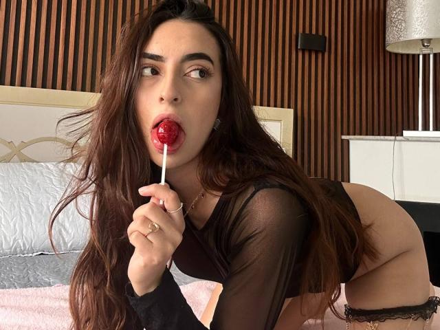 CelesteLarson with long brown hair, black sheer top, and red lollipop in her mouth, leaning forward, showing her ass in black lace panties, in a bedroom.