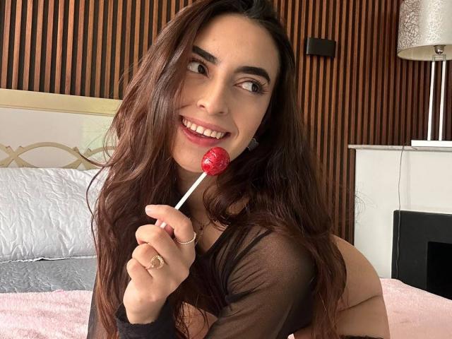 CelesteLarson, with long brown hair, holding a red lollipop, smiling, wearing a black sheer top, and showing her ass, leaning on a pink bed.