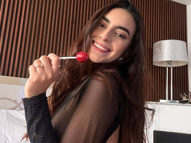 CelesteLarson with long brown hair, smiling, holding a red lollipop, wearing a sheer black top, showing her ring and white bed in the background.