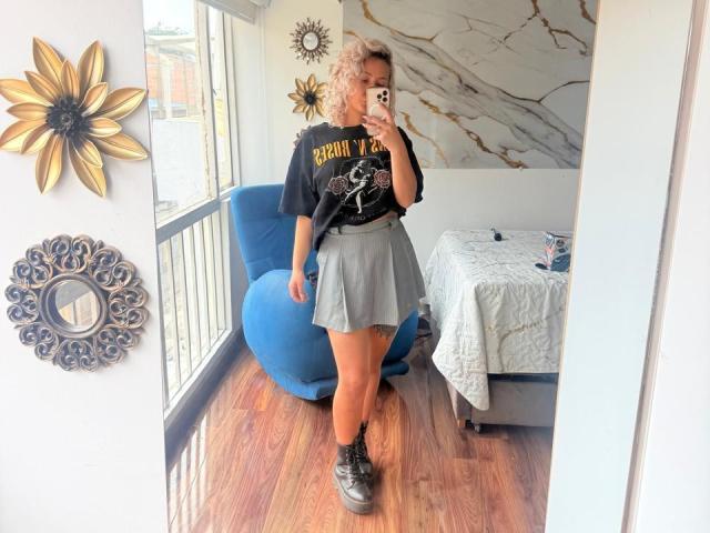 ElenaSaenz has curly blonde hair, a black "BRENDAN" t-shirt, gray skirt, black boots, and tan skin, standing in a room with a blue chair and wooden floor.