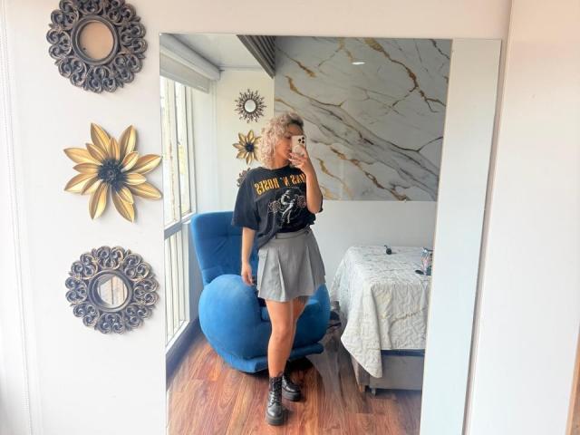 ElenaSaenz with curly blonde hair, black "BOOM YA!" shirt, gray skirt, and black sneakers, standing in front of a mirror, holding her phone, blue chair behind.