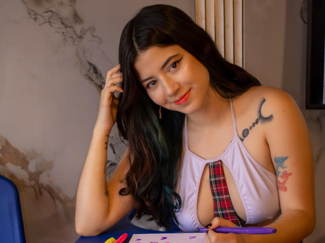 EmmaFeri with long dark hair, smiling, wearing a white halter top with a red plaid tie, exposing her tits, has tattoos on her arm and shoulder, writing at a table.