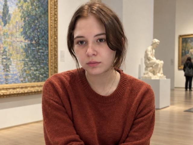 HoaGunby with brown hair, wearing a red sweater, has a neutral expression, sitting in an art gallery. Her ass is slightly visible under the sweater.