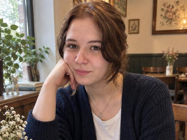 HoaGunby with brown wavy hair, wearing a navy cardigan and white tee, resting her chin on her hand, looking at the camera with a subtle smile.