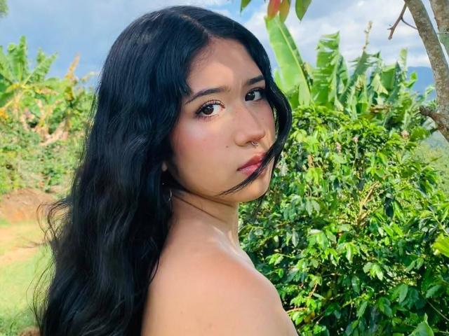 HannaThorne with long black wavy hair, nose ring, and serious expression, looking over her shoulder, topless, greenery and bananas in the background, outdoors, sunny day.
