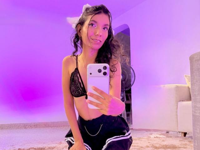 NyluLove met lang krullend haar, gekleed in een zwarte kanten bh en zwarte shorts, maakt een selfie in een paars verlichte kamer, terwijl ze een witte telefoon met drie camera's vasthoudt..
