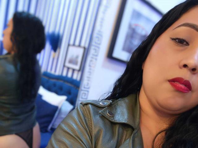 AmberMoretti with red lipstick, black hair, green leather jacket, facing camera, reflection shows her black panties, sitting on blue bed, looking over shoulder, watermark visible.