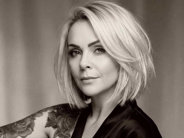 VictoriiaGreys with shoulder-length blonde hair, tattoo on left arm, wearing black satin top, looking at camera with slight smile, sepia-toned.