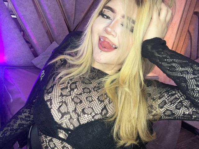 Lyanekkoo con i capelli biondi, indossa una maglia nera in pizzo, ha la lingua fuori, mostrando un piercing alla lingua, una mano tra i capelli, sorride, mostrando i suoi seni attraverso il pizzo, in una stanza poco illuminata..