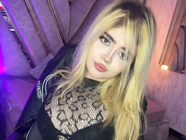 Lyanekkoo with blonde hair, black lace top, nose ring, and red lipstick, looking at the camera with a slight smile. Soft purple and pink lights in the background.