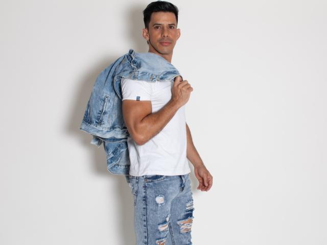 JoseRumBerotop with a lean, muscular build, short dark hair, wearing a white tee, ripped blue jeans, and a denim jacket over his shoulder, looking back with a slight smile.